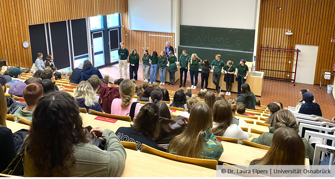 Blick von hinten in einen Hörsaal voller Studierender. Vorne stehen Mitglieder der Fachschaft und der Studiendekan Prof. Michael Hensel. An einer Tafel steht "Welcome".